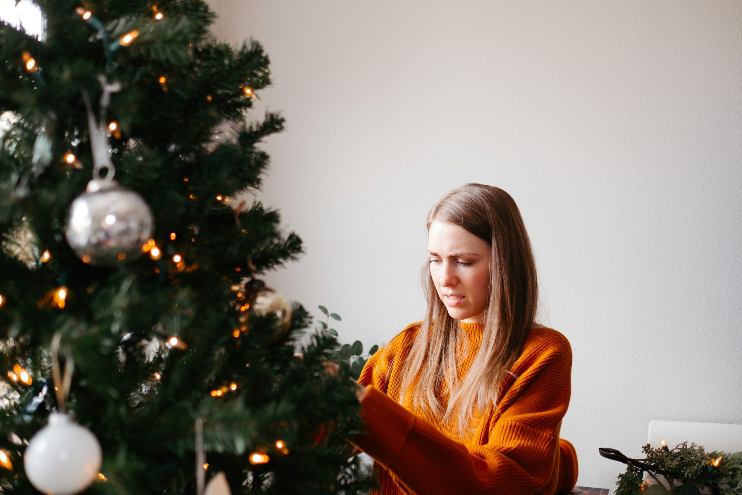 A woman in an orange sweater is joyfully decorating a Christmas tree with lights and ornaments, capturing the holiday spirit. The tree, partially visible on the left, features a prominent round silver bauble against a plain white wall backdrop.