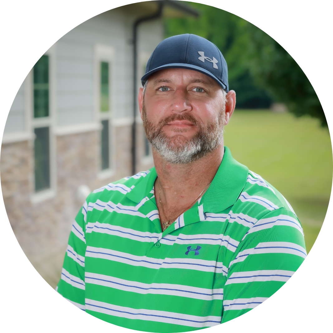 A man with a beard wearing a navy Under Armour cap and a green-striped polo shirt stands outside near a building with a lawn and trees in the background.