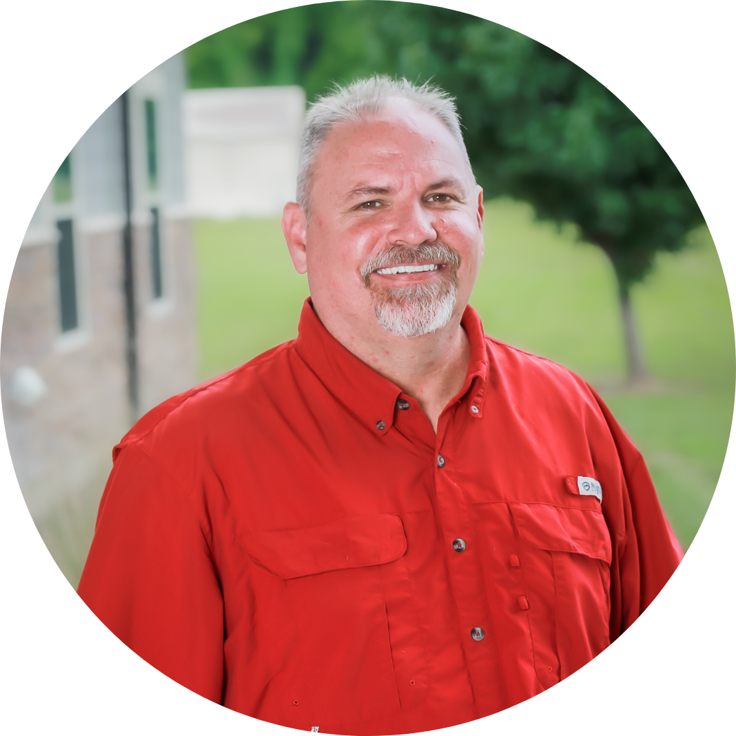 A smiling man with short gray hair and a goatee wears a bright red button-up shirt, standing outdoors in front of a building and greenery.