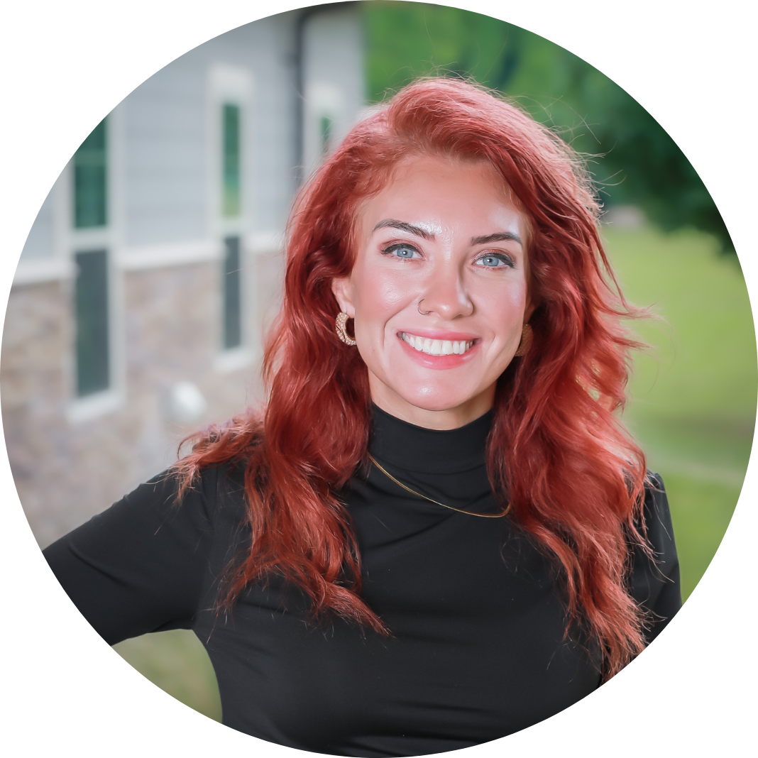 A woman with long, wavy red hair and blue eyes is smiling. She is wearing a black top and gold hoop earrings, standing outdoors with a blurred green background and a building in the distance.