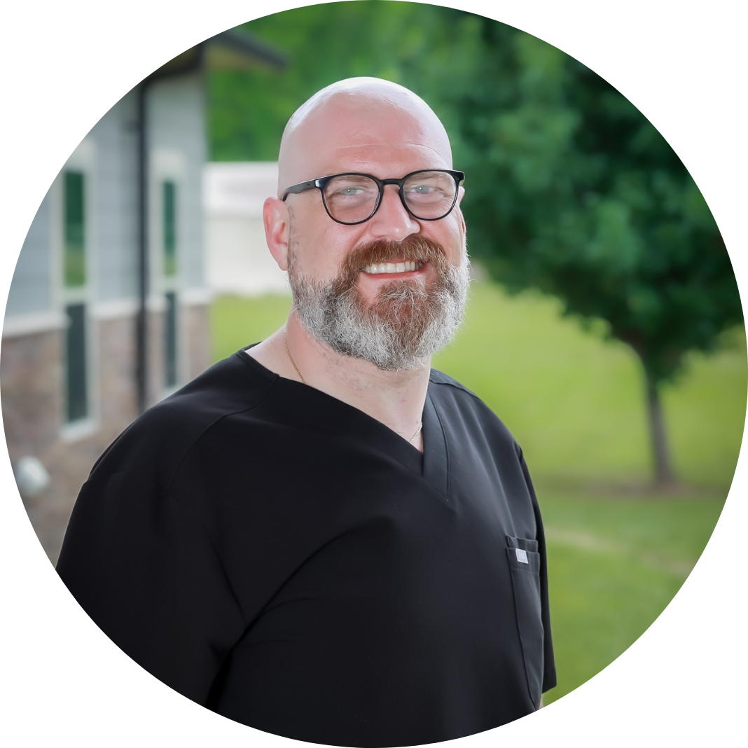 A smiling man with a bald head, glasses, and a beard, wearing black medical scrubs, stands outdoors near a building with grass and trees in the background.