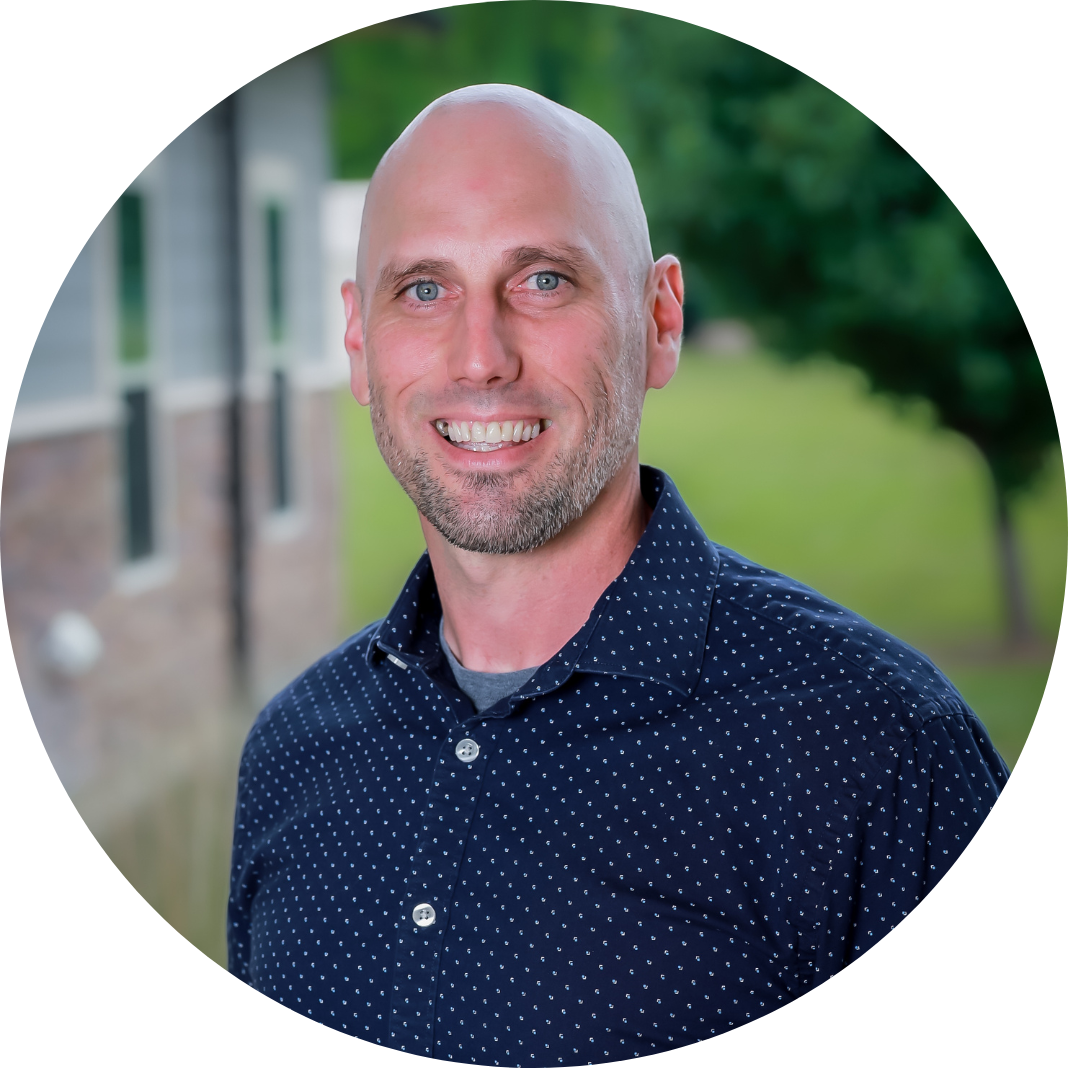 A smiling bald man with a trimmed beard wearing a navy blue shirt with white polka dots stands outdoors in front of a blurred background with greenery and a building.