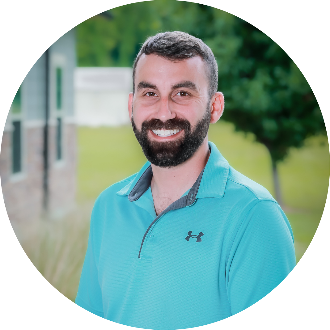 A man with a dark beard and short hair, wearing a teal Under Armour polo shirt, smiles outdoors with trees and a building in the blurred background.