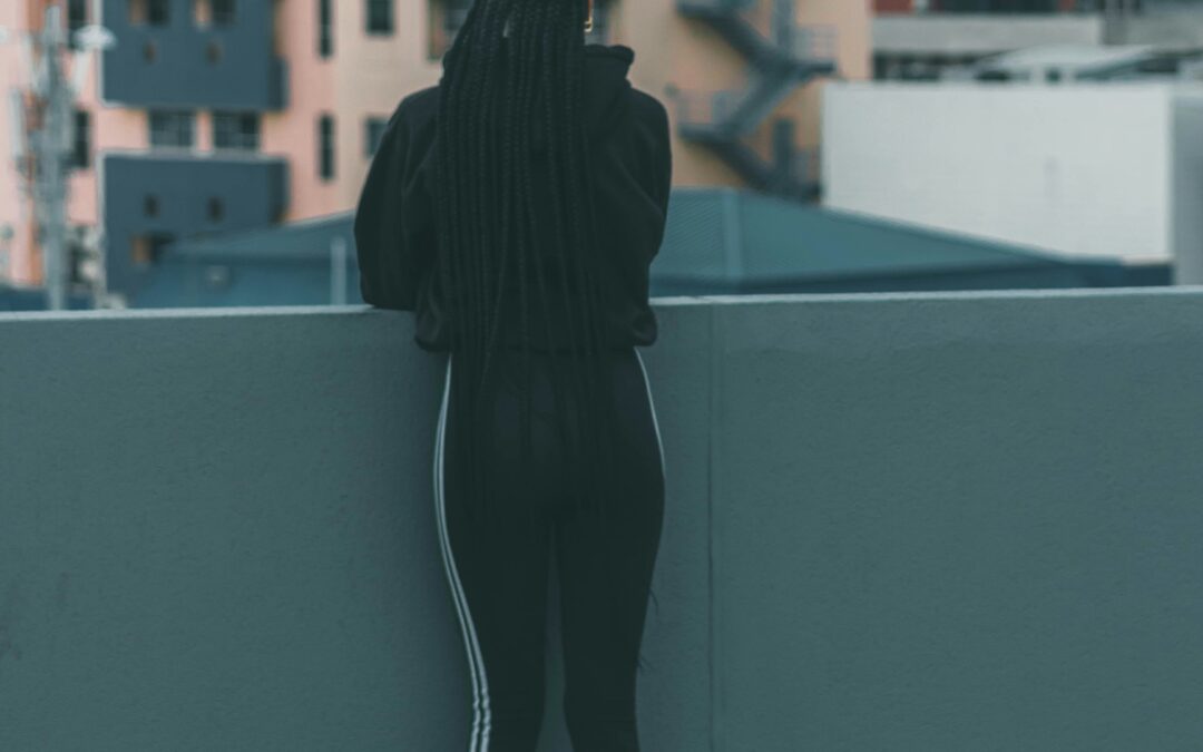 A person with long braided hair, dressed in black athletic wear, stands on a rooftop facing away, looking out over city buildings with a mountain in the background.