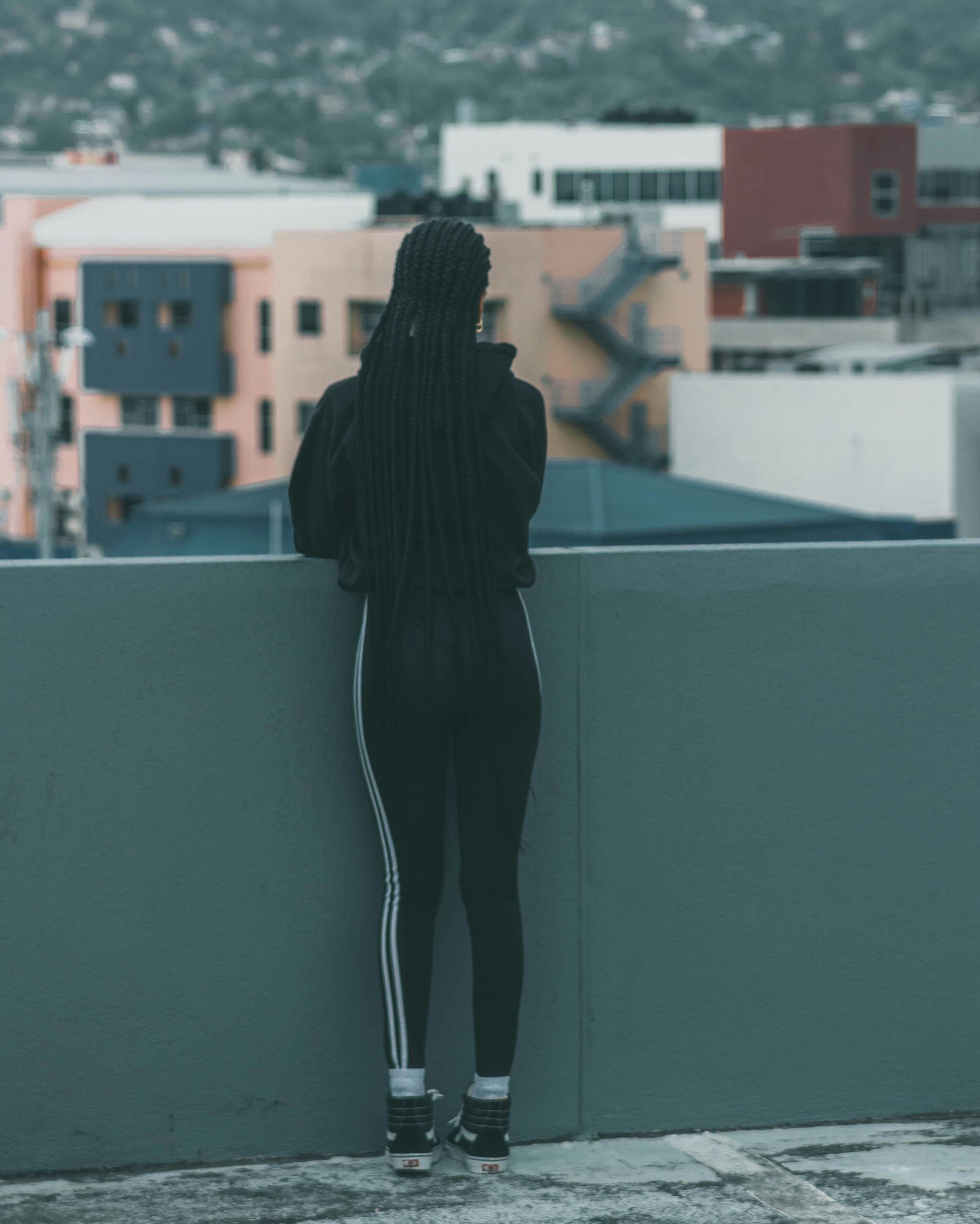 jabari-timothy-hPFXlHq5-As-unsplash A person with long braided hair, dressed in black athletic wear, stands on a rooftop facing away, looking out over city buildings with a mountain in the background.