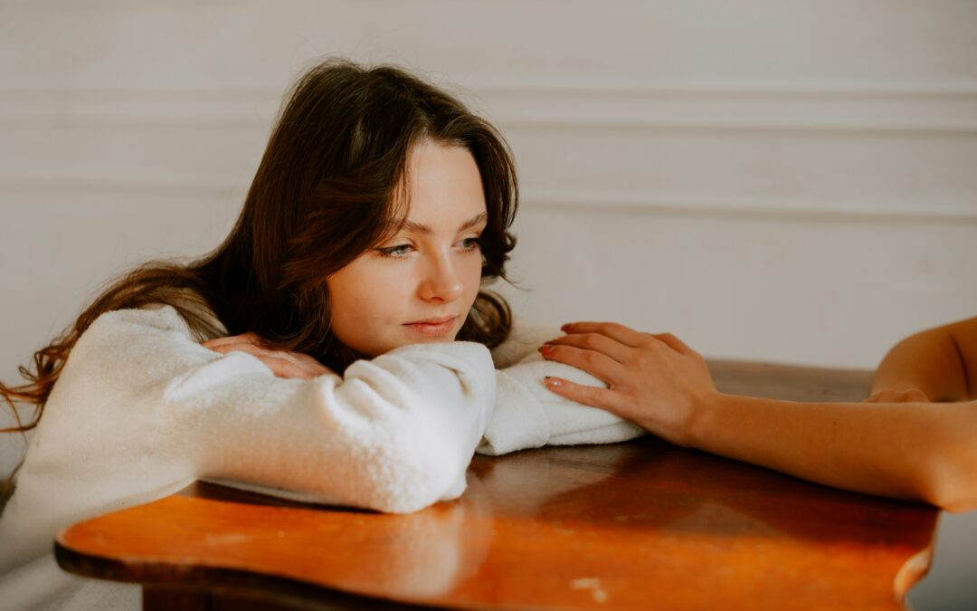 A young woman with long brown hair rests her chin on her crossed arms on a wooden table, looking thoughtful—much like someone weighing intensive outpatient vs inpatient choices. Another persons hand is gently placed near hers, offering comfort.