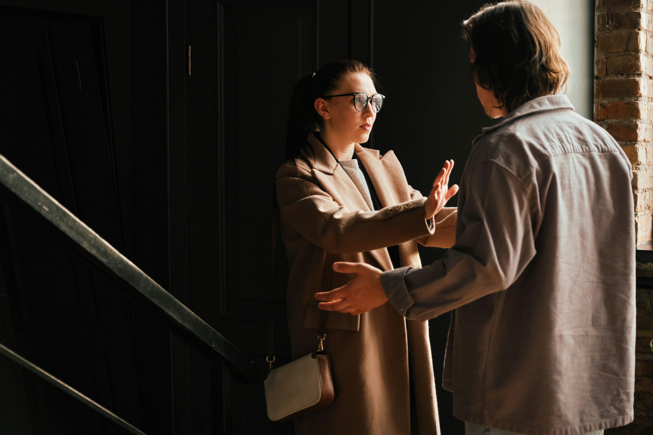andrej-lisakov-lvI5utYLY4E-unsplash A woman in a brown coat raises her hands to gesture stop while speaking to a man in a gray shirt indoors, near a staircase and brick wall, both appearing to have a serious conversation about holistic therapy.
