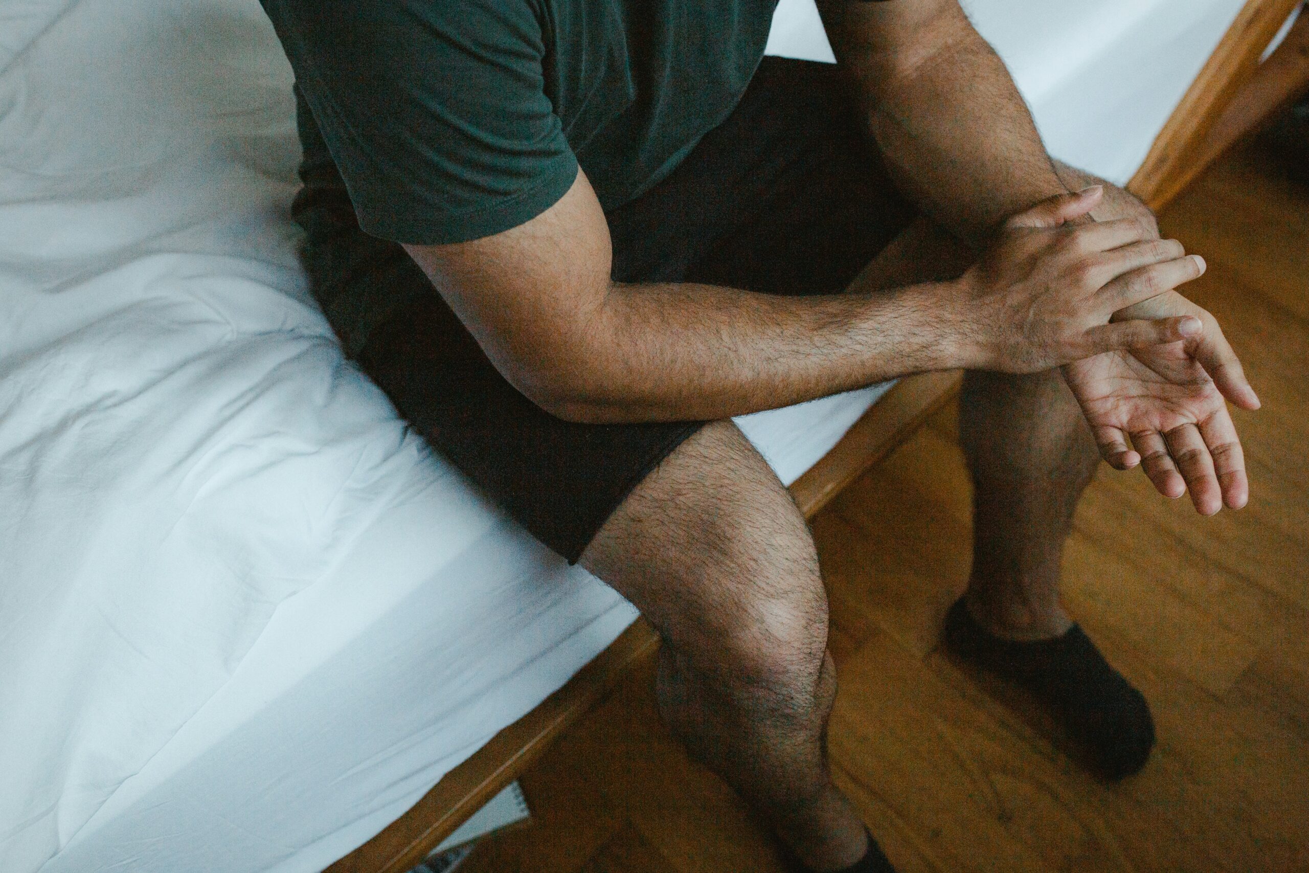 victoria-romulo-XXmJIp2Apwg-unsplash A person wearing a dark green shirt, black shorts, and black socks sits on the edge of a bed, with hands resting on their knees in a relaxed posture. The image is cropped to show only the torso and legs.