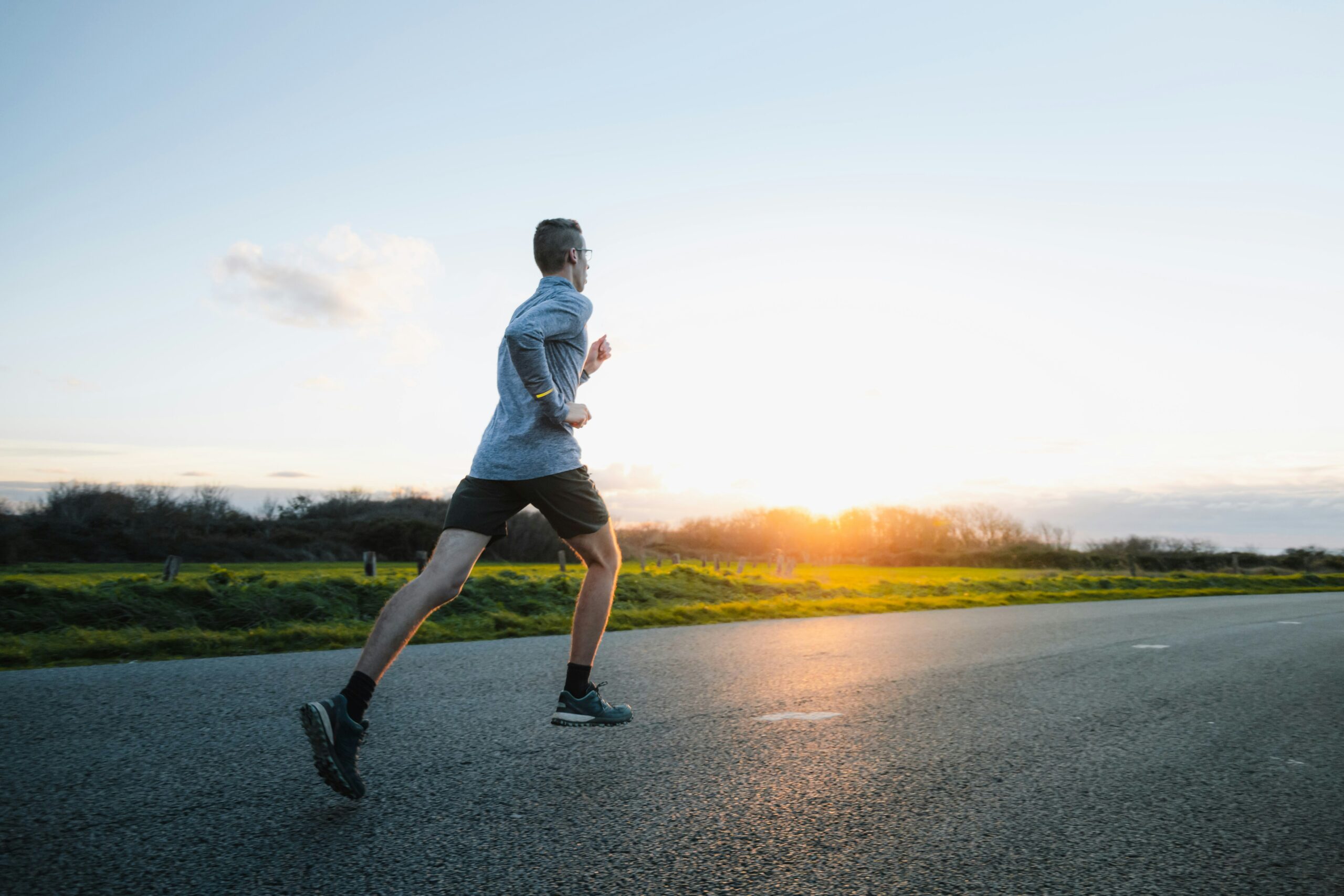 benoit-deschasaux-2oOtH40fF90-unsplash A person wearing a gray long-sleeve shirt and black shorts runs along an empty road at sunrise, with green grass and bushes on either side under a clear sky.