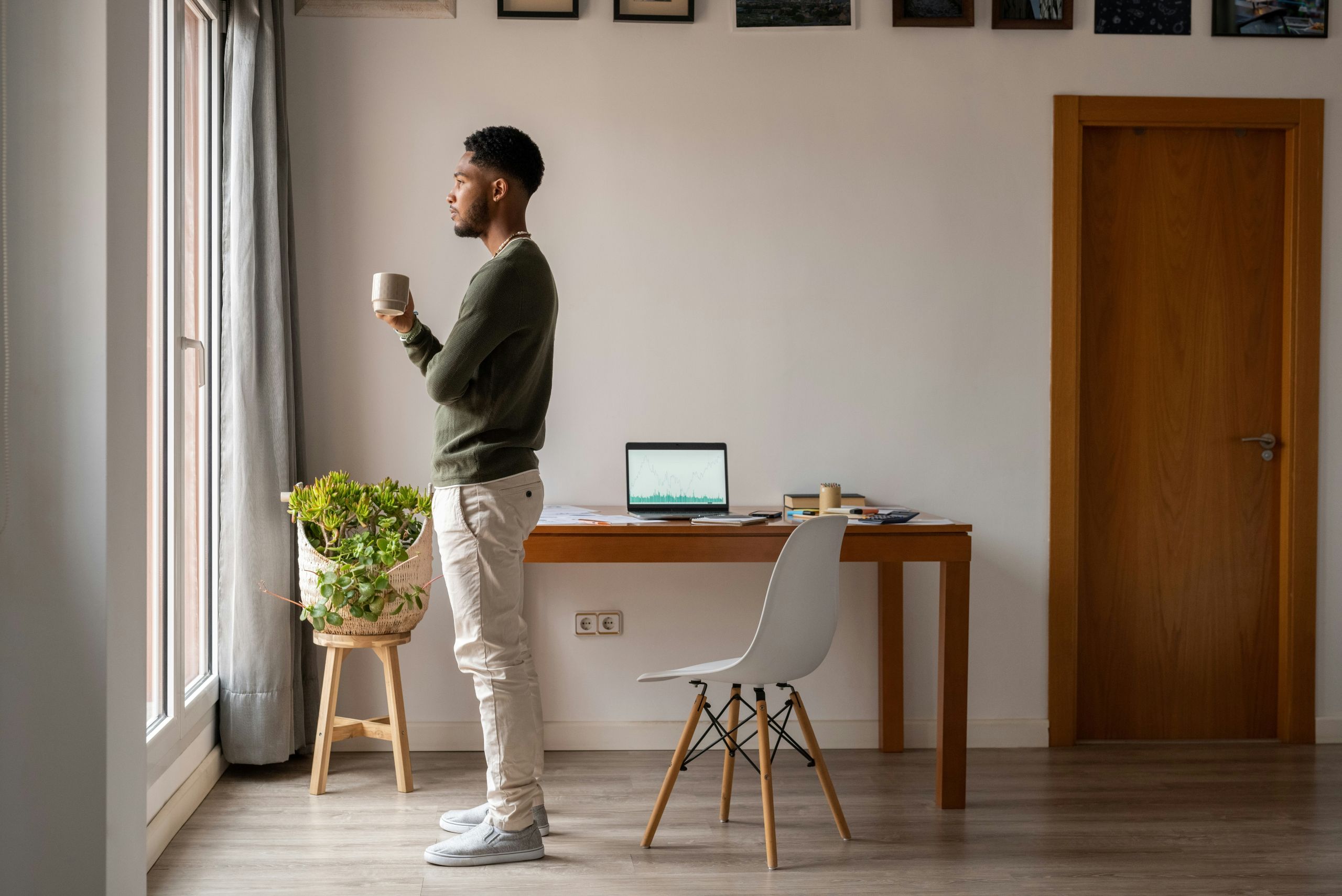 A man stands by a window holding a mug, looking outside thoughtfully. Behind him is a wooden desk with a laptop, plant, and chair in a modern, tidy home office.
