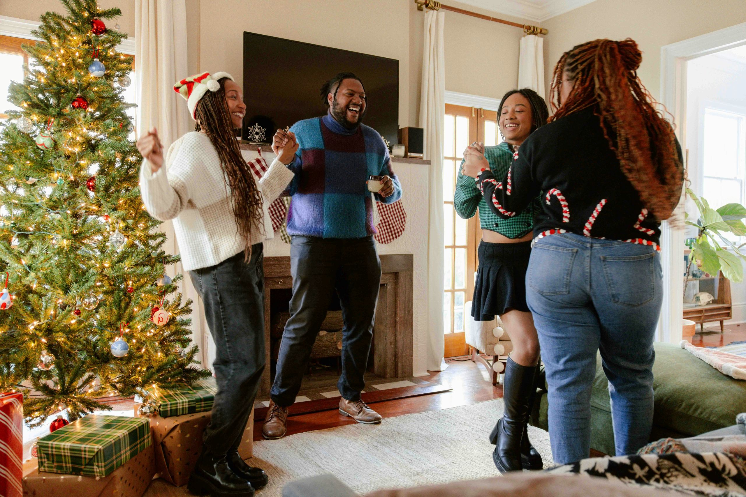 Four people joyfully dance and laugh together in a cozy living room decorated for Christmas, navigating family dynamics with warmth amid a brightly lit tree, presents, and a glowing fireplace in the background.