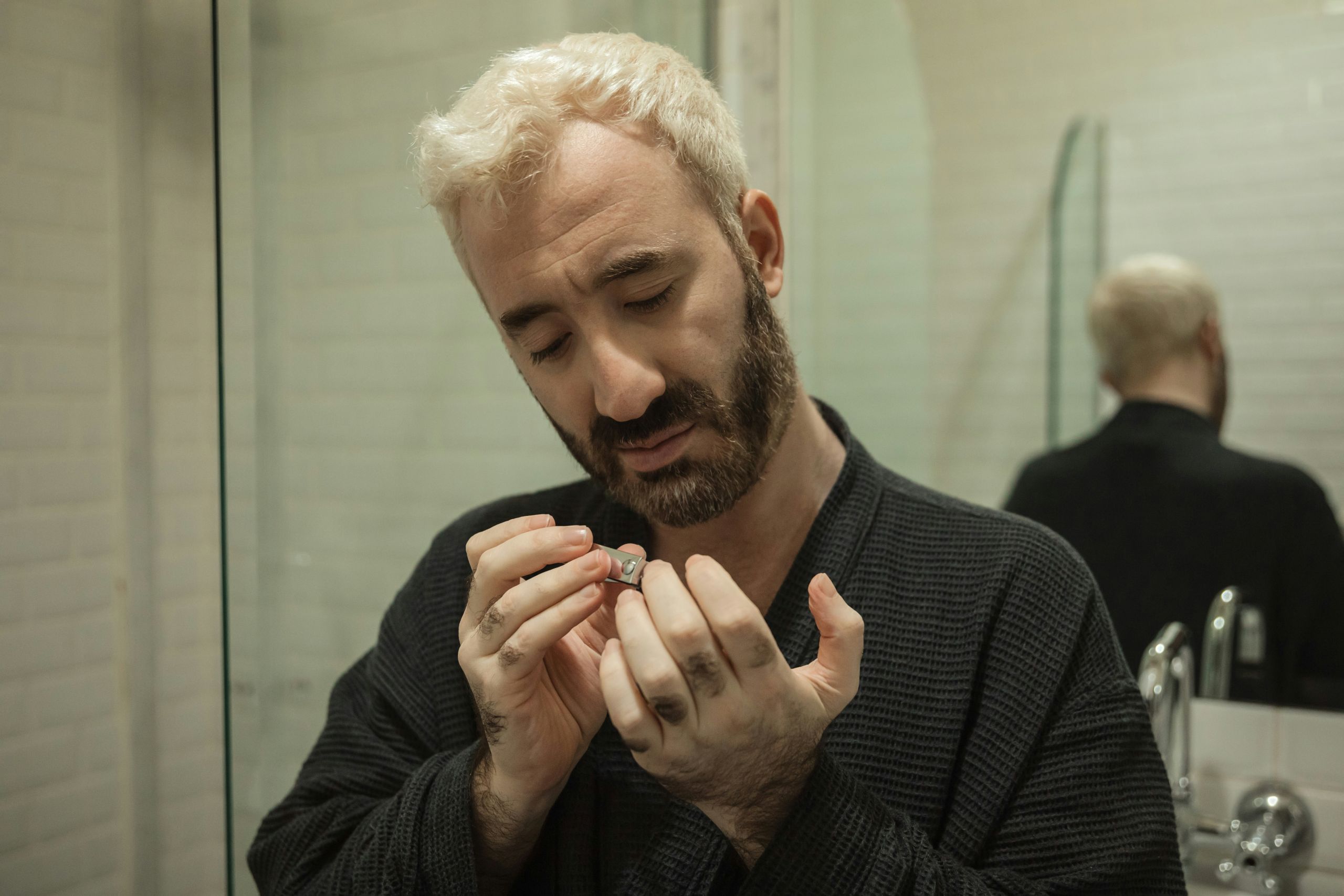 A man with bleached blond hair and a beard, wearing a dark robe, stands in a bathroom and files his fingernails—a quiet moment of self-care in recovery, his reflection visible in the mirror behind him.
