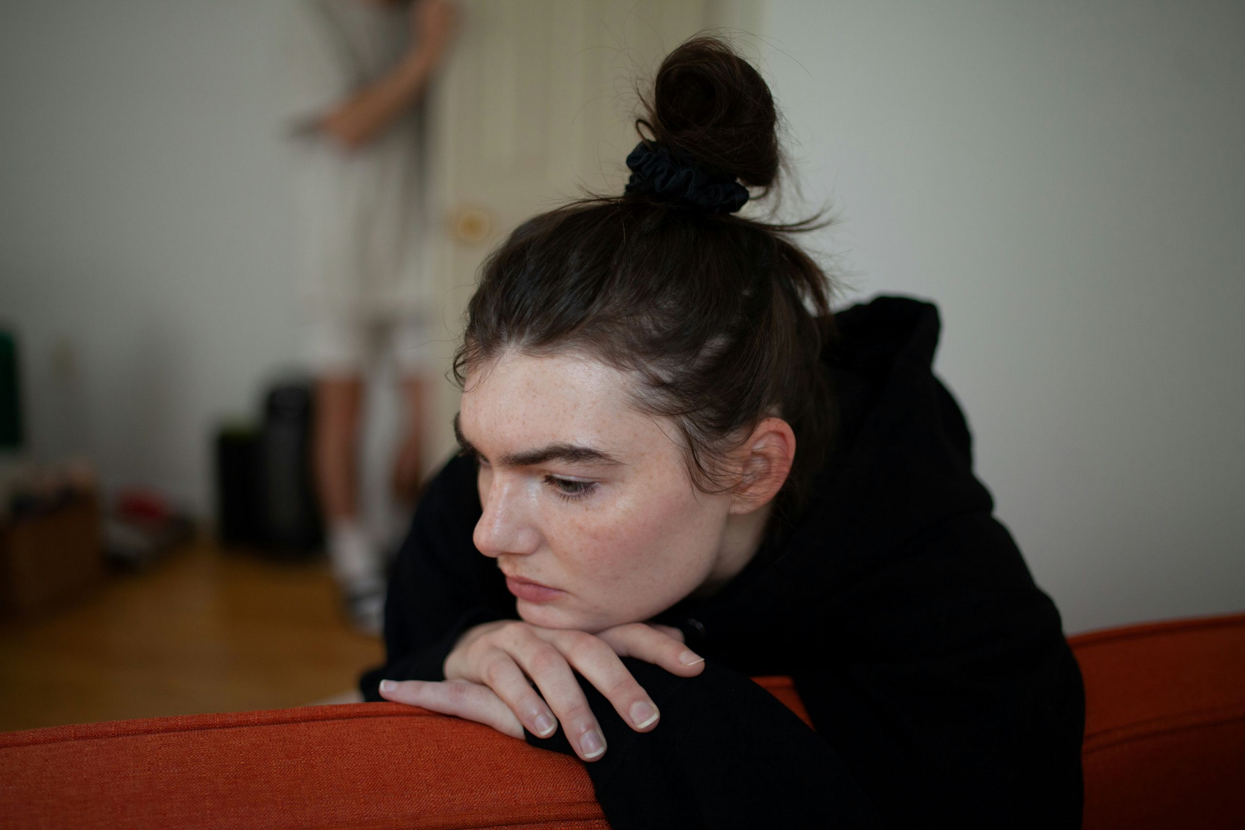 A person with dark hair in a messy bun leans forward on the back of an orange couch, looking down thoughtfully, as if reflecting on emotional healing. The background shows a blurred figure standing and a white door.