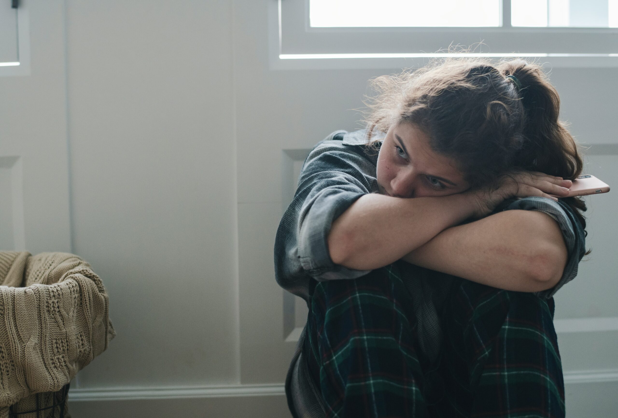 A woman sits on the floor indoors, hugging her knees and resting her head on her arms with a pensive expression, possibly reflecting a seasonal mood shift. She wears plaid pants and a gray shirt as natural light streams through the window behind her.