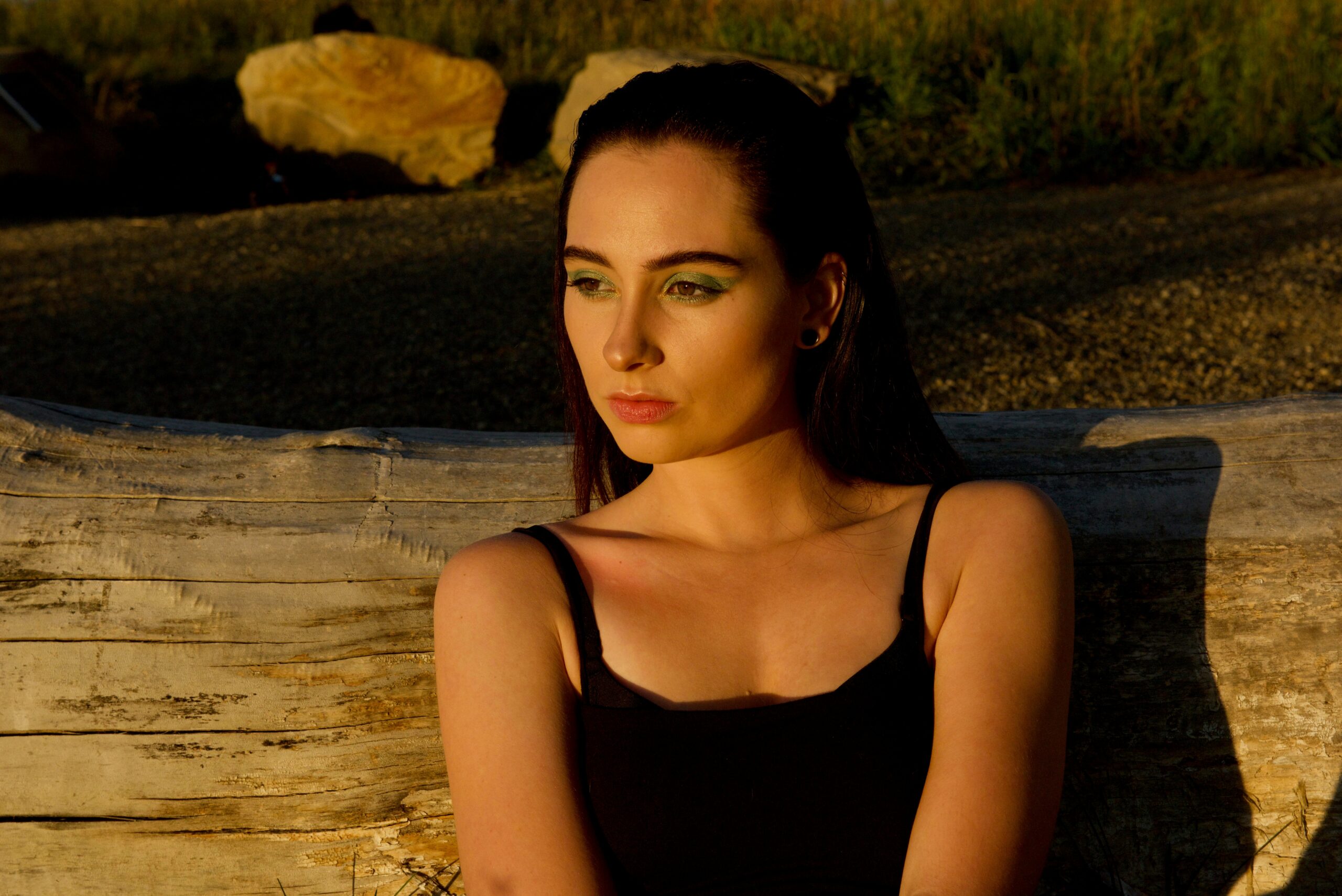 A woman with long dark hair and green eye makeup sits on a large log outdoors, bathed in warm sunlight, looking thoughtfully to the side as if reflecting on trigger management. She is wearing a black tank top.