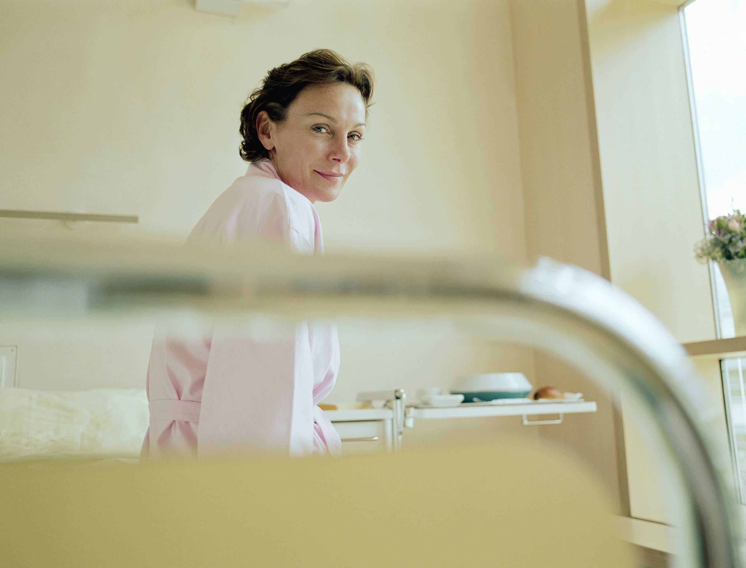 A woman wearing a pink robe sits on a hospital bed, looking over her shoulder and smiling slightly. The bright room, filled with natural light and a vase of flowers on the windowsill, reflects the supportive environment of dual diagnosis treatment.