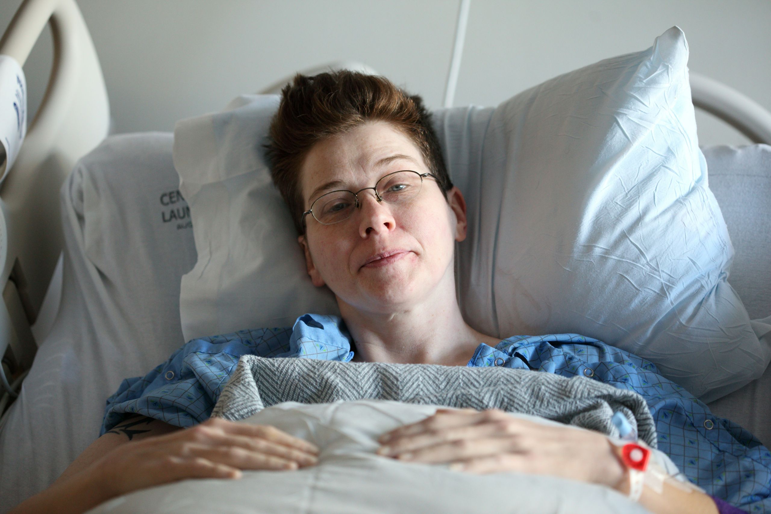 A person with short hair and glasses lies in a hospital bed, wearing a hospital gown and resting under a blanket, looking at the camera with a faint smile while receiving medication-assisted treatment.