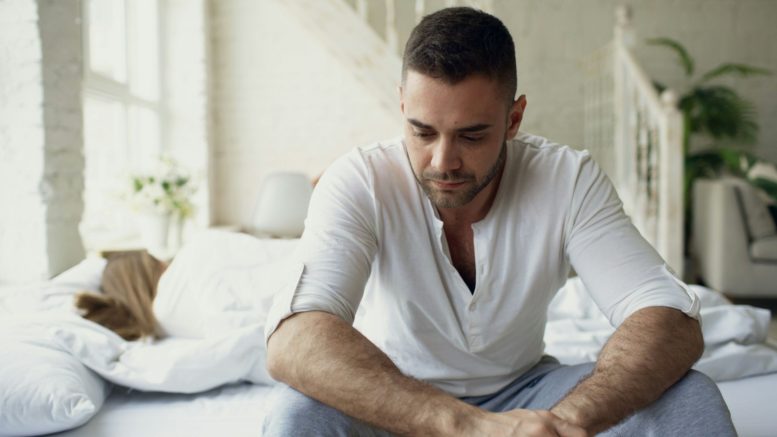 addiction treatment A man sits on the edge of a bed with his head slightly bowed, looking thoughtful or worried—possibly reflecting on addiction treatment. The room is bright, with white bedding and soft natural light. Another person lies in bed, mostly out of focus.