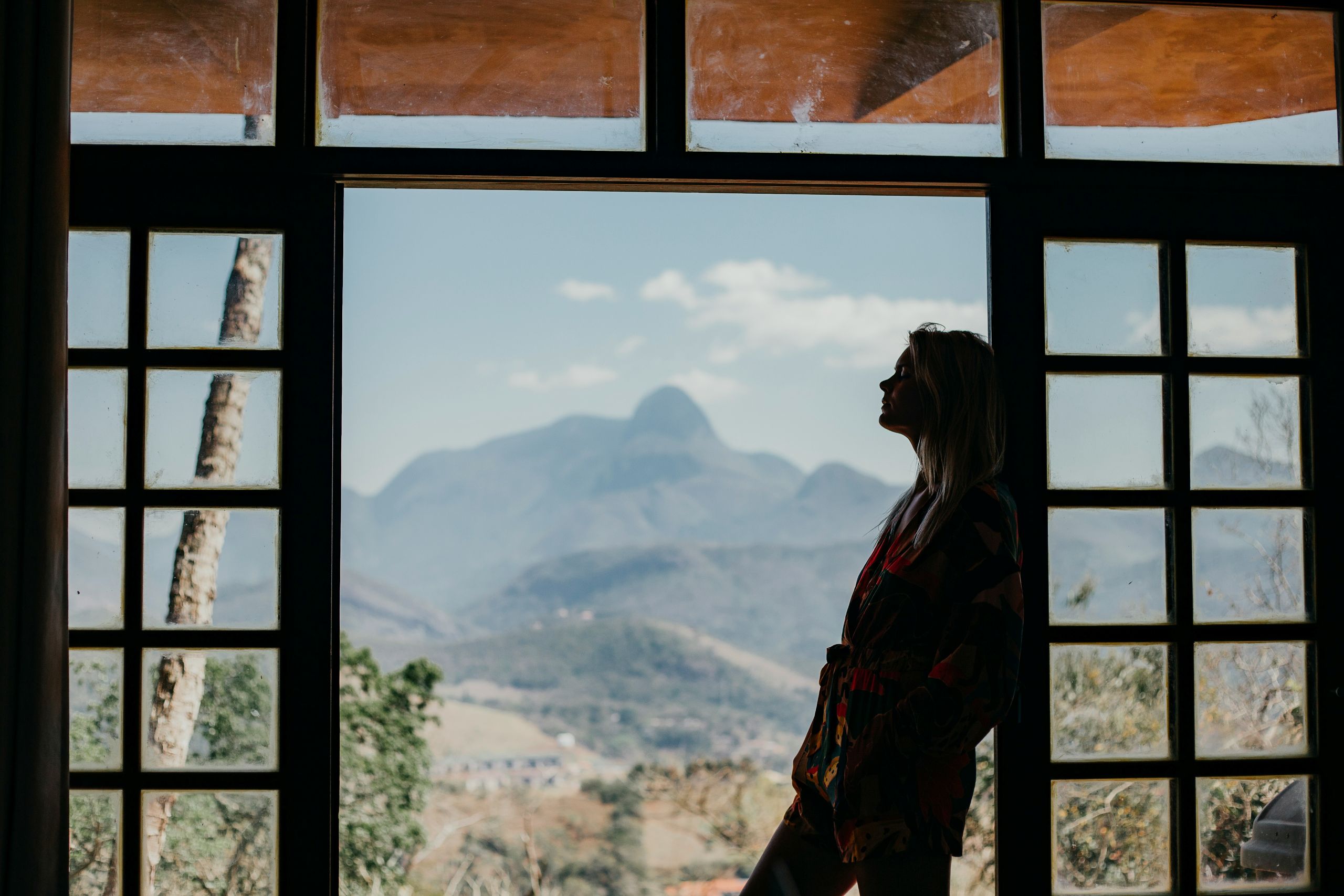 structured addiction programs A woman stands in silhouette by large window panes, looking out at scenic mountains under a partly cloudy sky. Natural light fills the room, highlighting the peaceful, contemplative atmosphere often found in structured addiction programs.