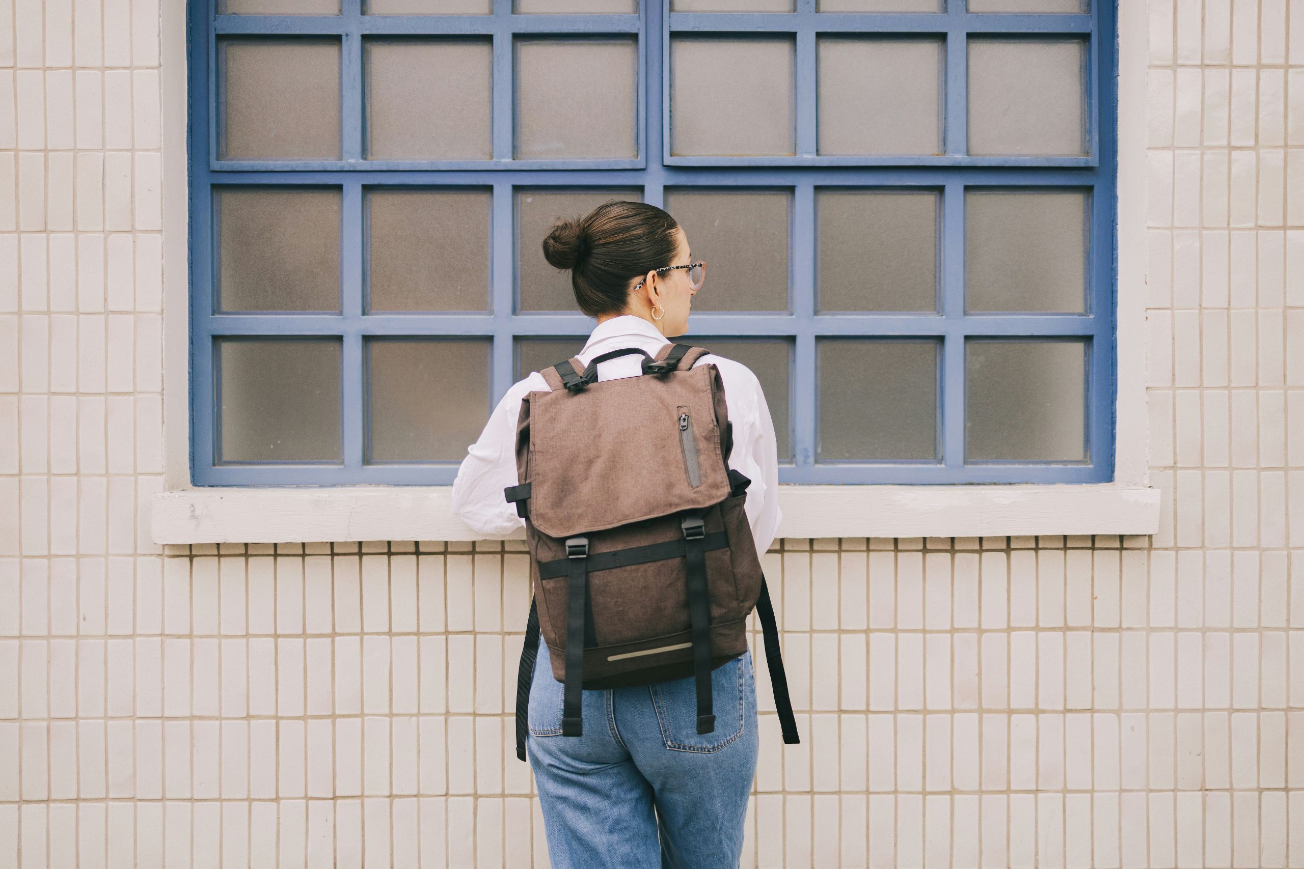 sobriety A person with a bun hairstyle, wearing glasses, a white shirt, jeans, and a brown backpack stands in quiet sobriety facing a frosted glass window with blue frames on a tiled wall.
