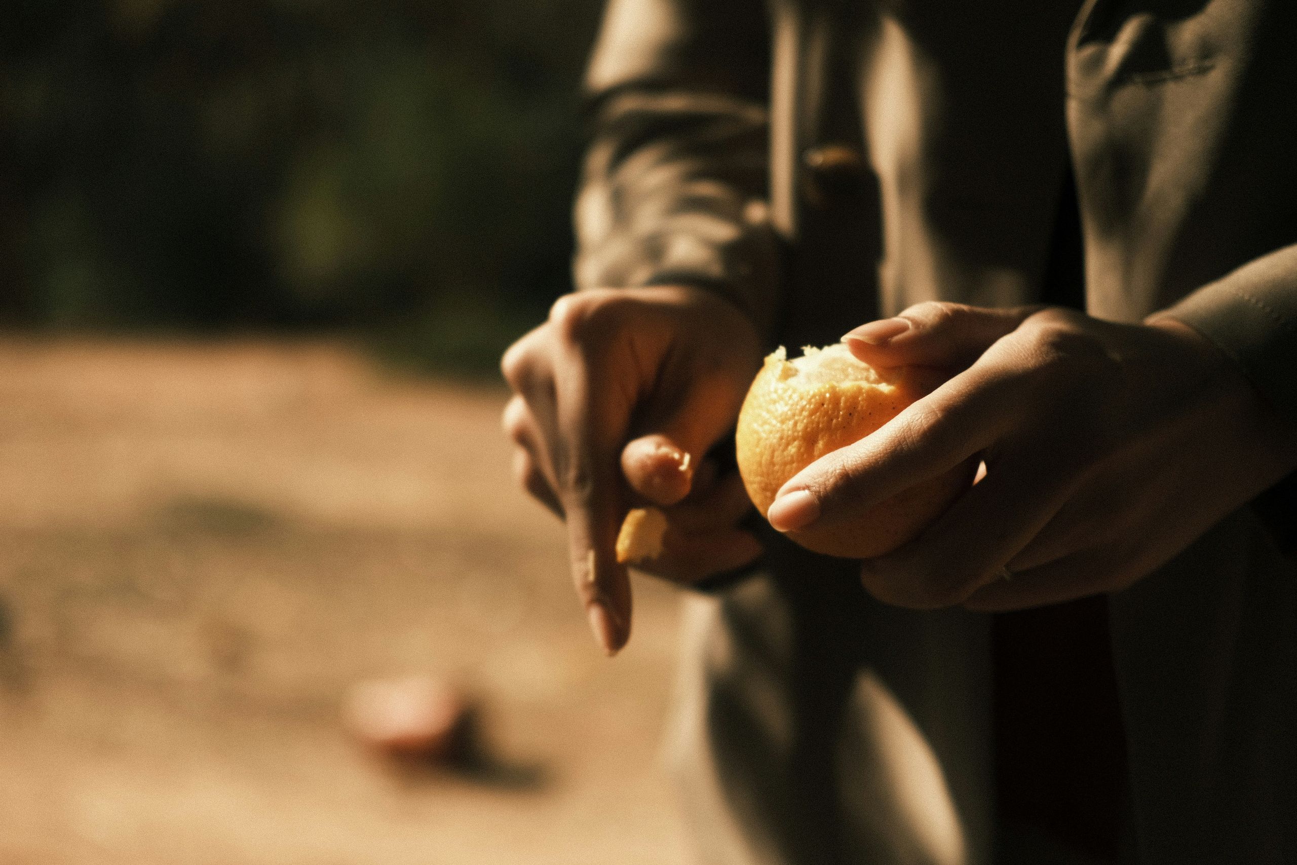 A person peels an orange with their hands outdoors, with soft sunlight highlighting their fingers and the fruit. The background is blurred, making the focus on the orange and the persons hands.