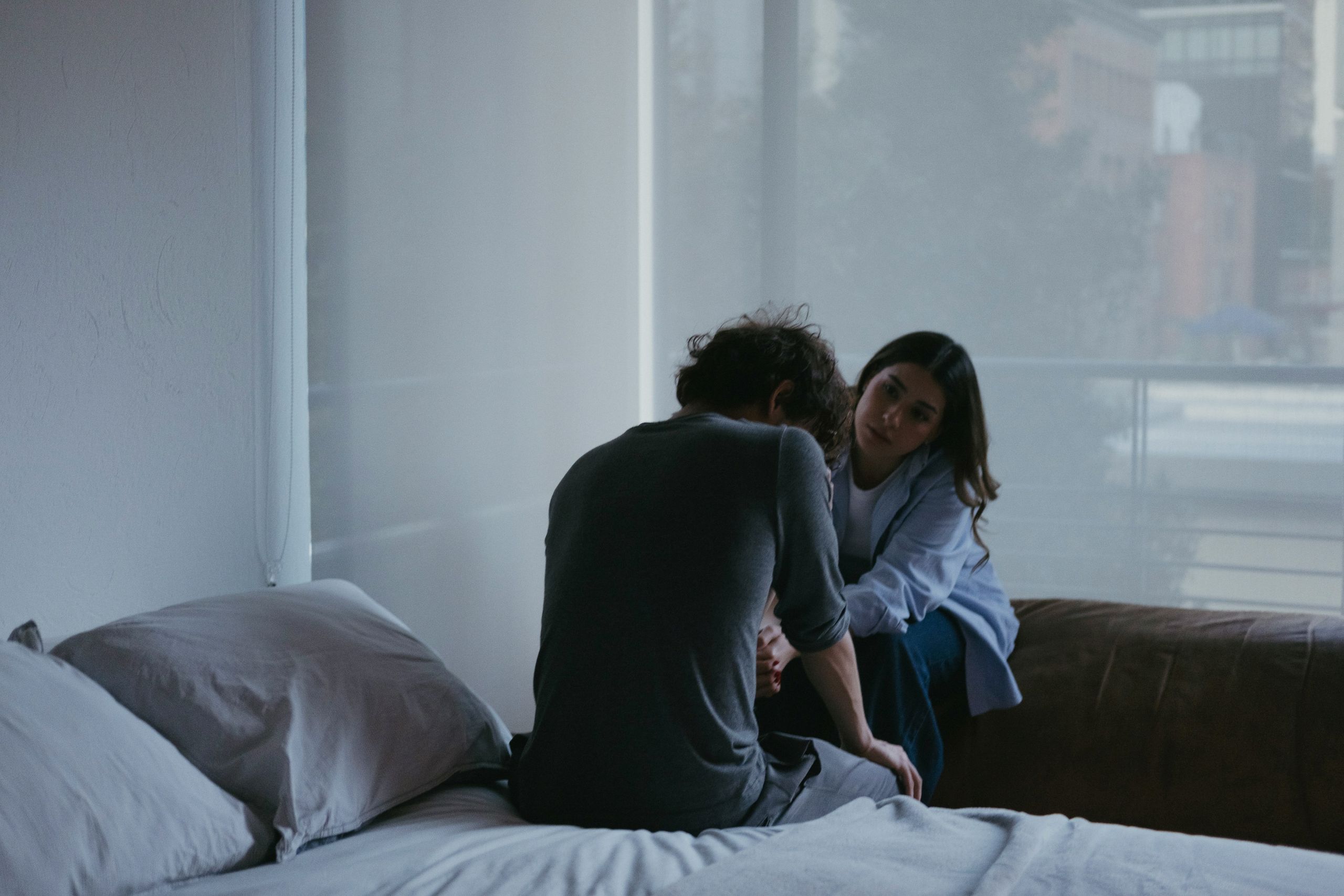 How to Build Healthy Friendships in Recovery A person sits on a bed with their back turned, while another person sits nearby, gently holding their arm and looking at them with concern—offering quiet support during recovery. The room is softly lit, with large windows in the background.