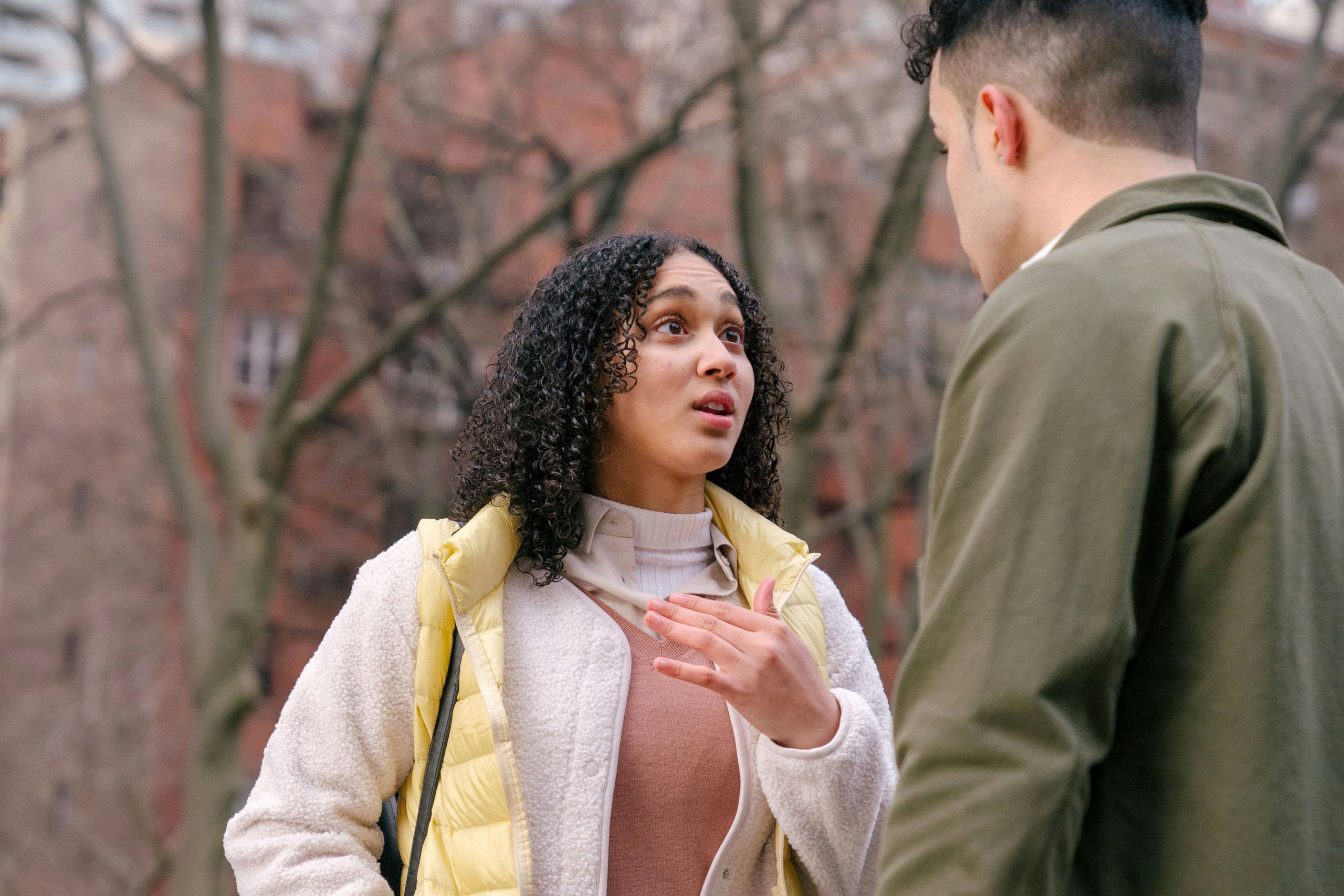 Why Honesty Is One of the Most Important Parts of Recovery A woman in a yellow vest speaks emotionally to a man in an olive jacket outdoors, their conversation shaped by honesty in recovery. Bare trees and a brick building form the backdrop to this heartfelt exchange.
