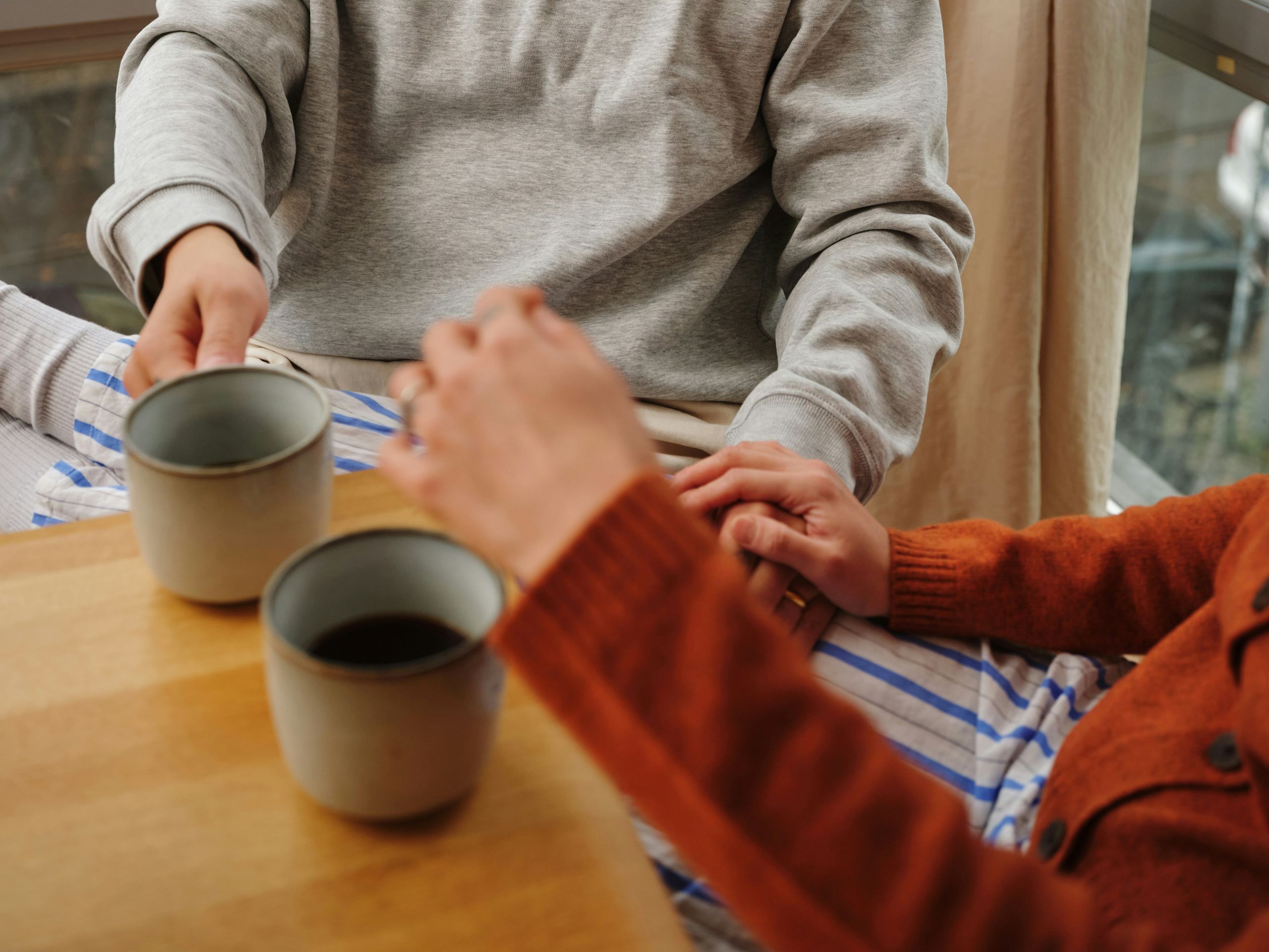 Two people sit closely at a table with two mugs of coffee, holding hands. One wears a gray sweater, and the other wears an orange sweater. Sunlight streams through a window beside them.