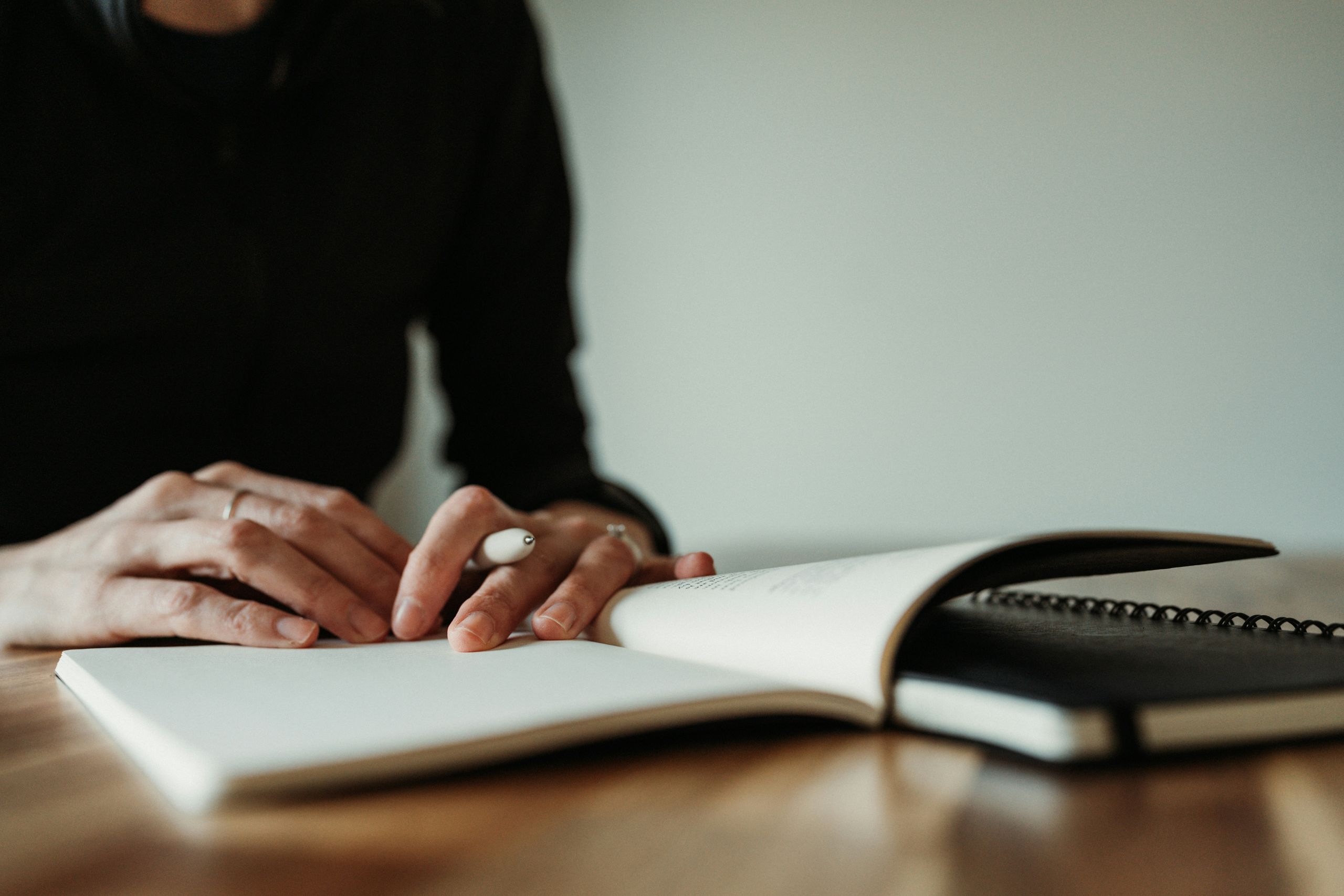 stress at work A person sits at a wooden table, holding a pen and writing or preparing to write in an open notebook, with another closed notebook underneath.