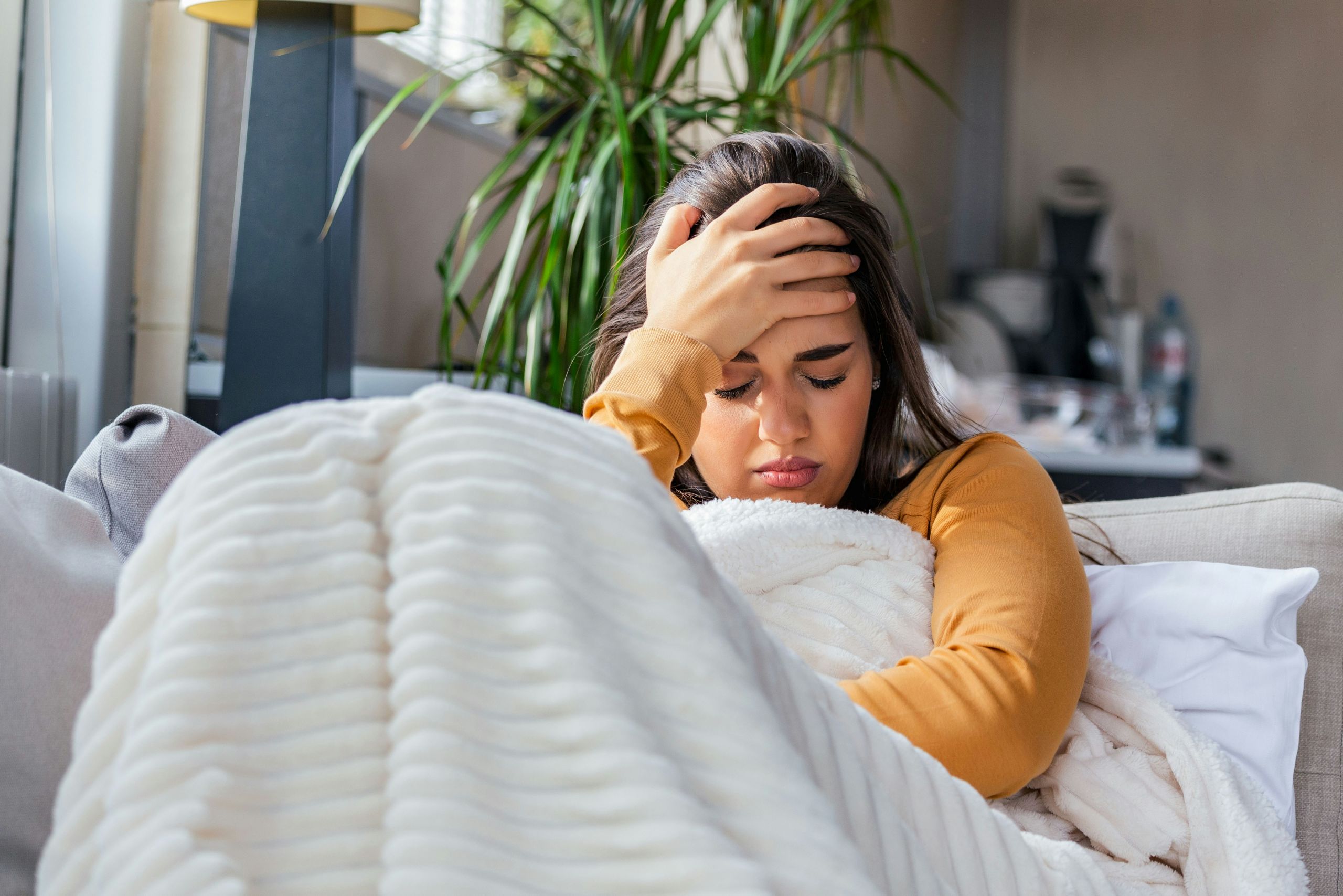 A young woman with a pained expression sits on a sofa, holding her head with one hand while wrapped in a white blanket.